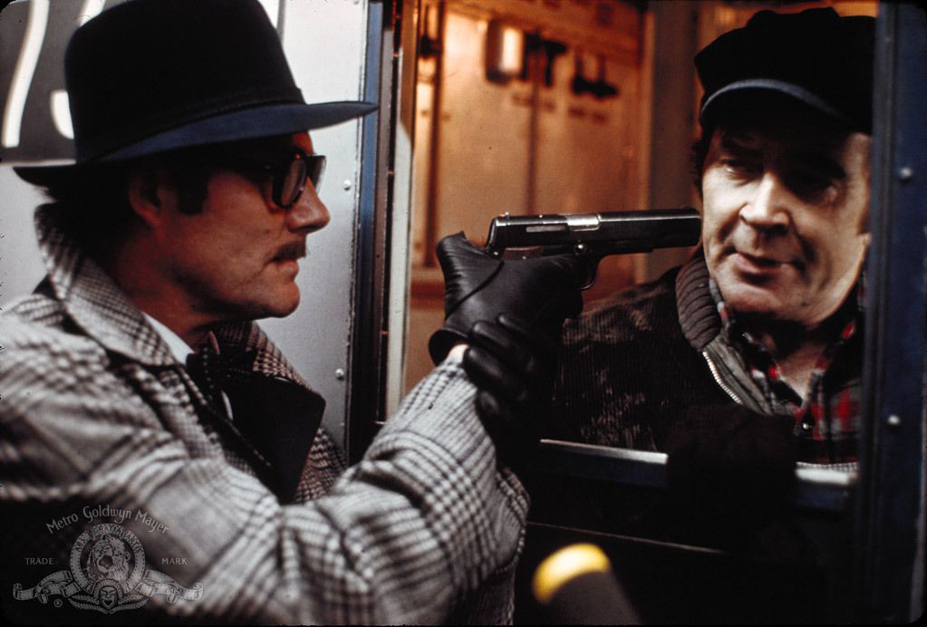 A man holds a gun through the open window of a train, pointed at another man. 