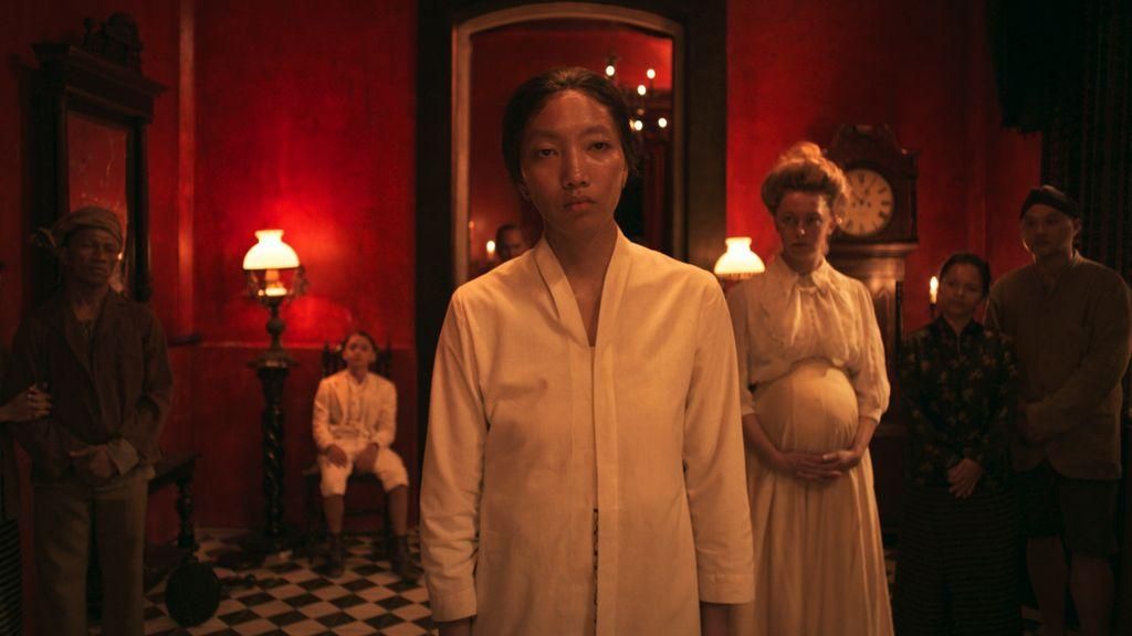 A woman in a white cotton robe stands in a red lit, old fashioned room, other people in white robes and worker's clothes stand behind her. 