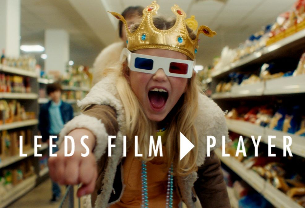A still from a short film with a child sat in a shopping trolley, with a toy crown and 3D glasses. The text says "Leeds Film Player"