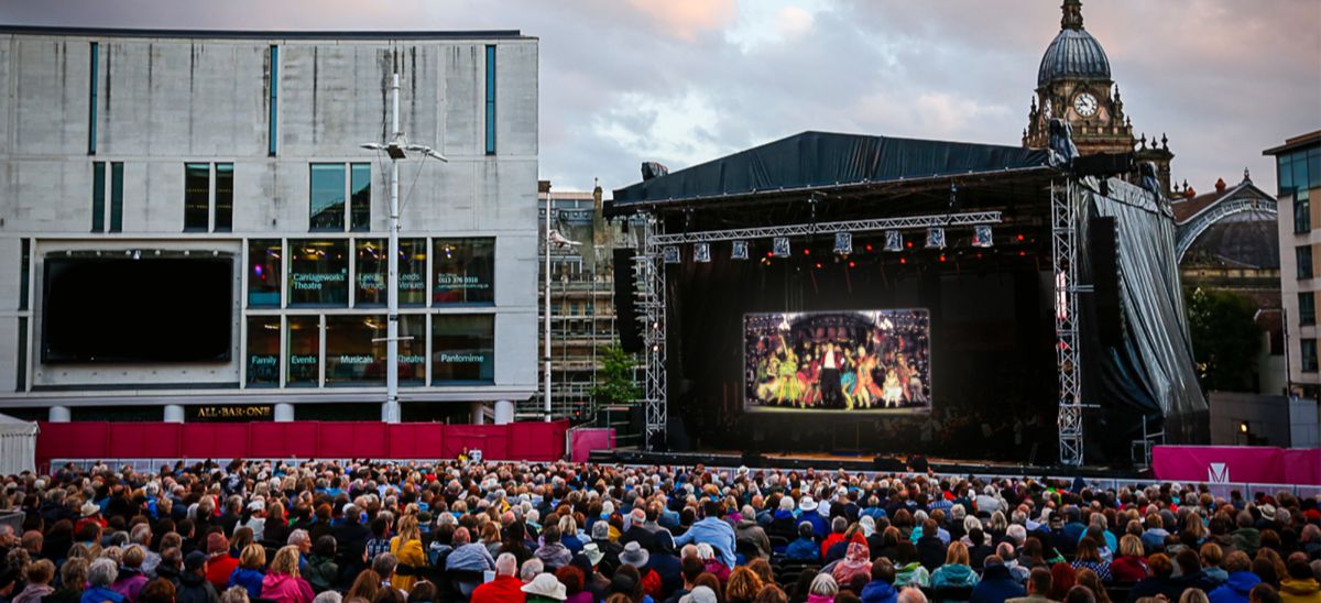 A large outdoor concert stage, lit up in the evening with a large audience watching the stage.