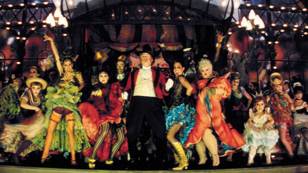 A stage full of colourfully dressed woman in frilly dresses gather around a man in a circus ringleader outfit.