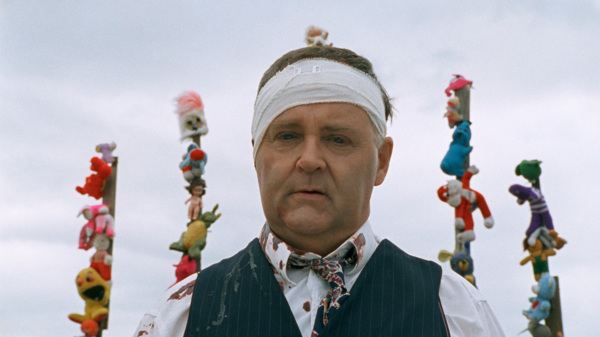 Still from Body Melt, showing a man with a bandage around his head, standing in front of poles covered in stuffed animal toys.