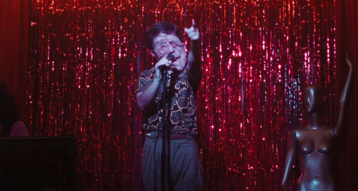 A man with a facial deformity confidently sings into a microphone on a small stage with red glittery streamers behind him. He points to the audience as he sings. 