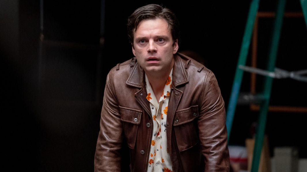 A man played by Sebastian Stan, in a brown leather jacket and floral shirt looks perplexed. 