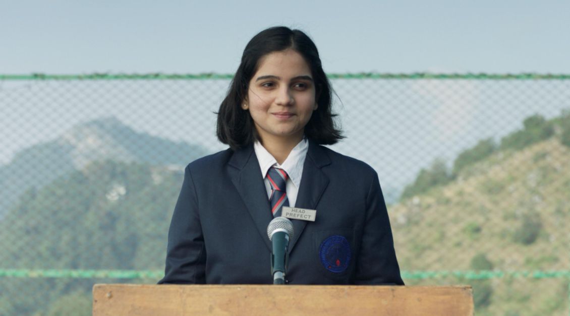 A 16 year old girl stands on an outdoor podium with a head prefect badge, making a speech. 