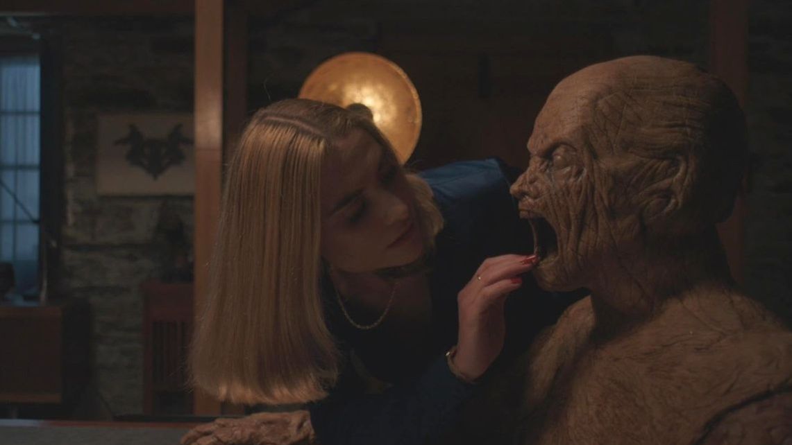 A blonde woman reaches her hand towards the open mouth of a sinister looking wooden mannequin. 