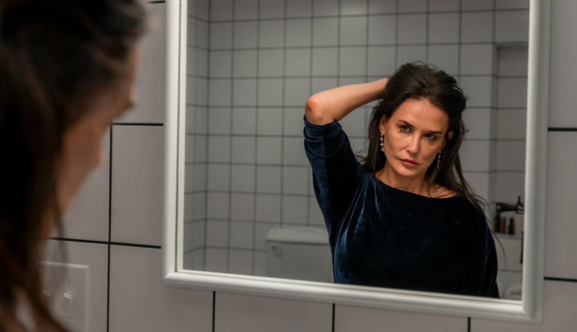 A older woman (played by Demi Moore) strands in from of a mirror, eyeliner smudged, posing and holding her hair up. 