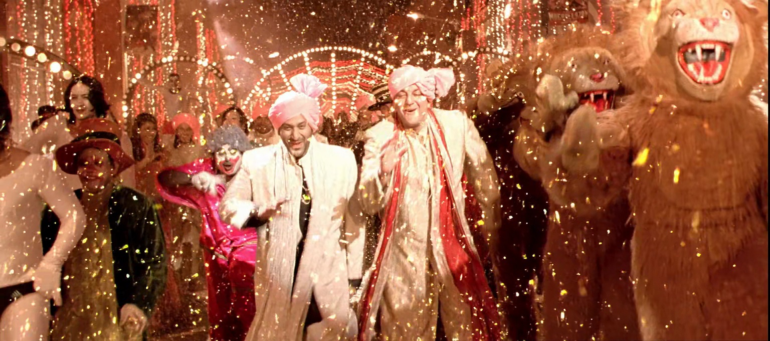A lively Bollywood scene from a film, with men in pink turbans and sherwanis dancing joyfully among sparkles and people in festive costumes.
