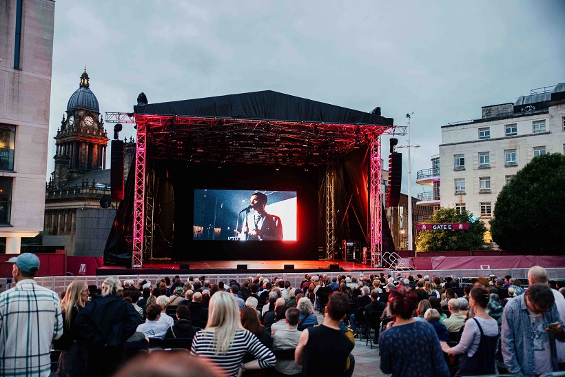 A large audience watches an outdoor film screening in Millennium Square, with Leeds Town Hall and surrounding buildings visible in the background.