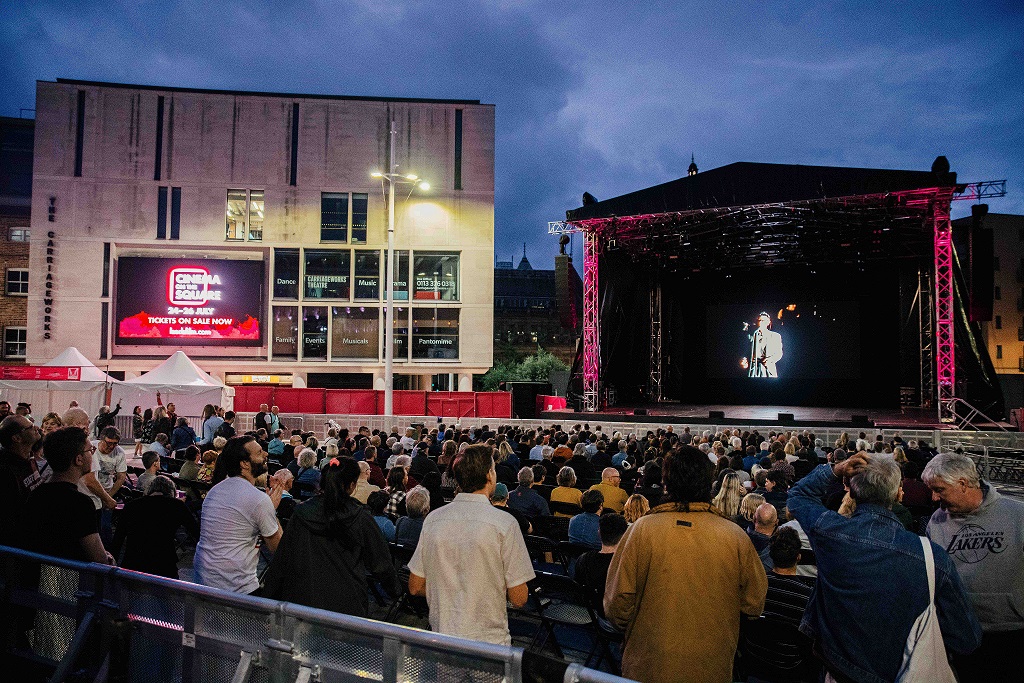 A large outdoor crowd gathered in front of a stage at dusk, watching a film screening at Cinema on the Square, with The Carriageworks building in the background.