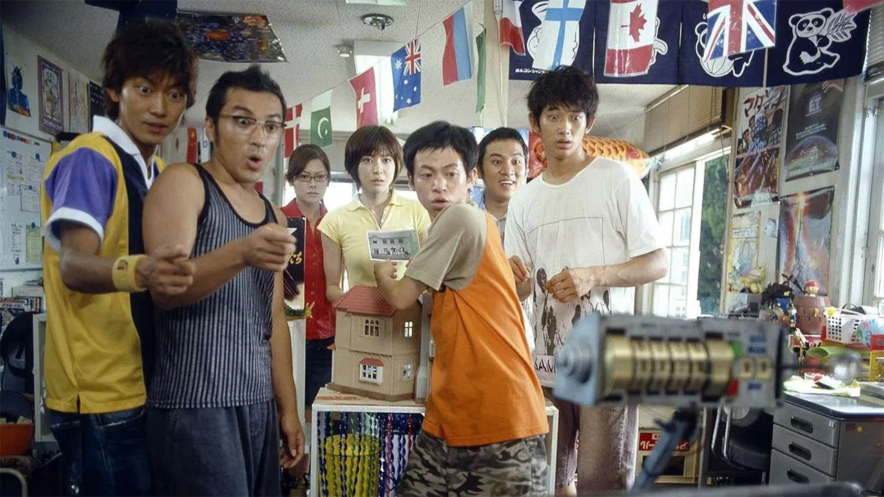 A group of surprised young men and women in a cluttered room, looking toward a strange mechanical object, in a still from the Japanese sci-fi comedy Summer Time Machine Blues.