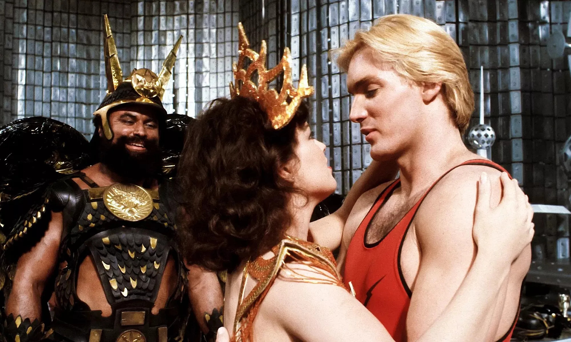 A fantasy film scene with a man in a gold crown embracing a woman in gold and red armour, while a smiling man in ornate black and gold armour stands behind them.