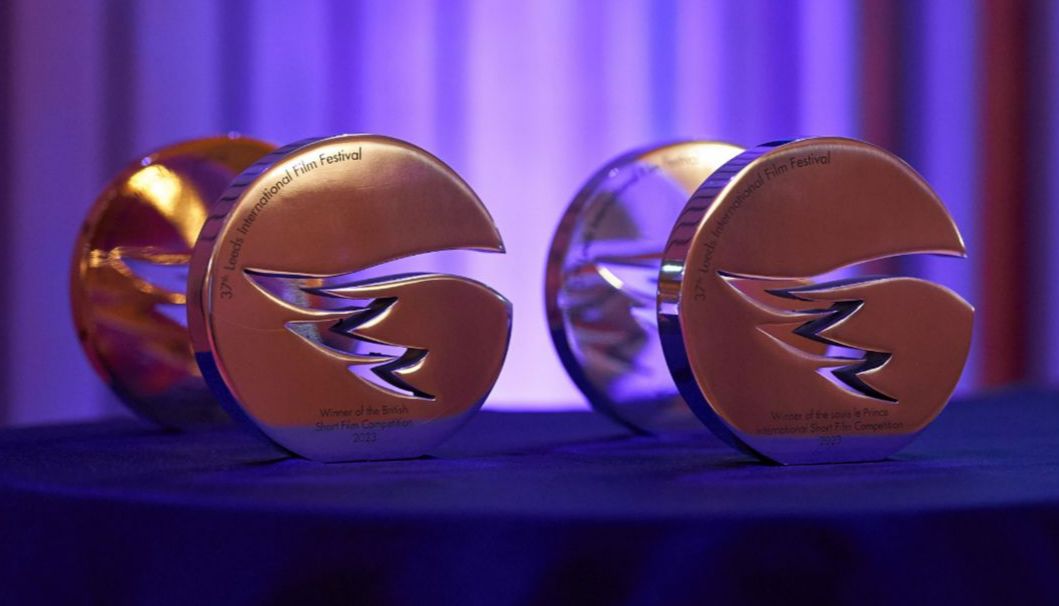 Two metallic Leeds International Film Festival awards rest on a dark tablecloth, illuminated by purple and blue lighting, engraved with the festival name and competition titles.