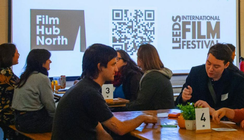 People sit at long tables in front of a large screen displaying Film Hub North and Leeds International Film Festival logos during a networking or industry event.