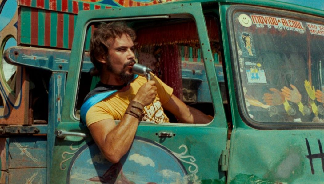 A man with a beard and long hair sits in a brightly painted van, holding a microphone to his mouth, surrounded by colourful patterns and stickers.