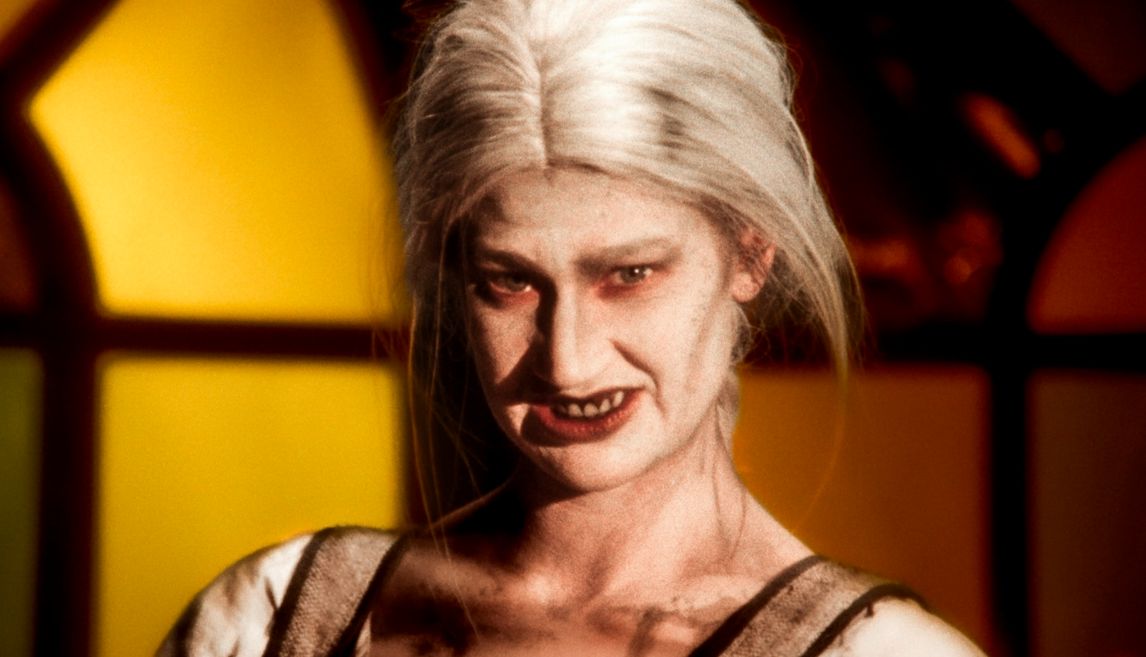 A pale woman with long white hair and dark makeup gives a sinister grin, showing sharp teeth, lit by warm light from stained glass windows behind her.
