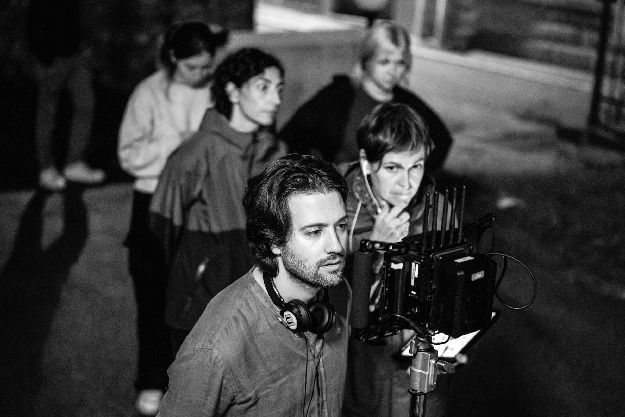 A black-and-white photo showing a film director and crew focused on a monitor during a shoot, the director wearing headphones and looking intently at the screen.