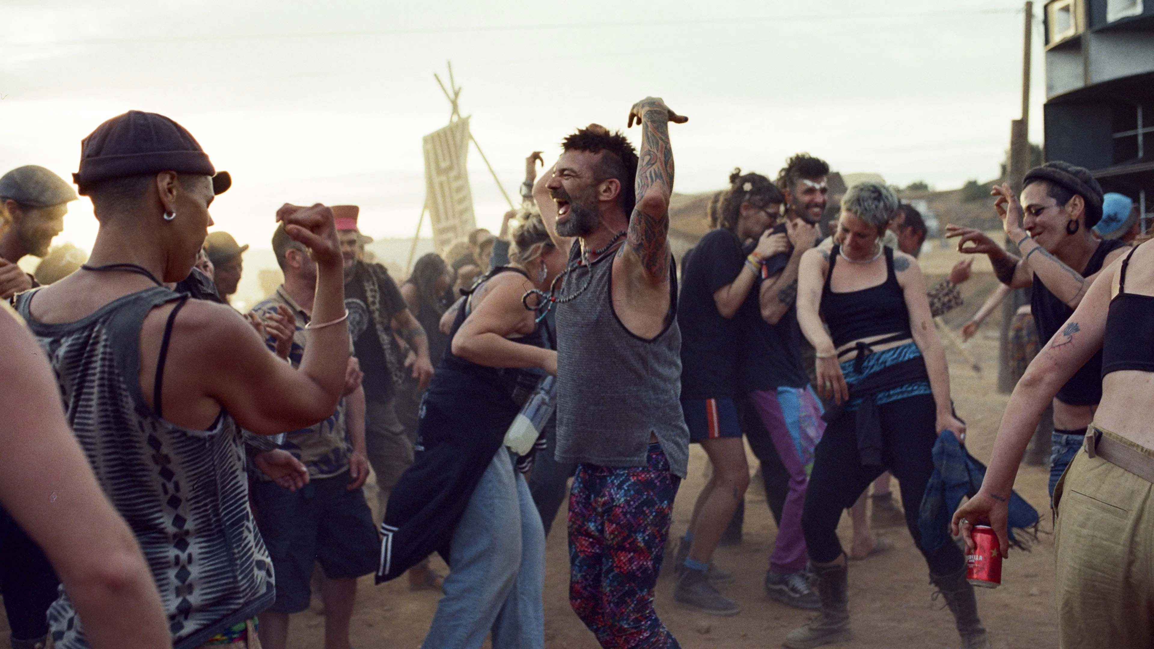 A group of people dancing outdoors at sunset, covered in dust and dressed in colourful clothes, celebrating with laughter and movement at what looks like a festival or rave.
