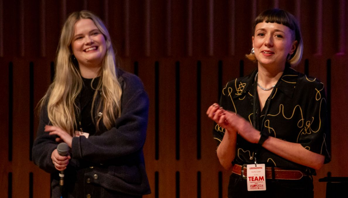 Two smiling women stand on stage at a film event, one holding a microphone, the other applauding, both wearing LIFF staff badges.