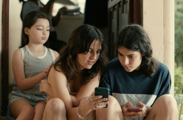 Two teenagers are looking at something on a mobile phone as a younger child sits behind them, playing with the older girls hair. 