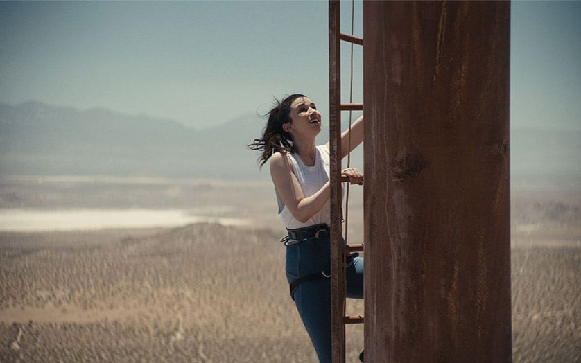 A young woman climbs up an old ladder of a rusted radio tower, high above the ground with a sprawling desert and mountain range behind her. 