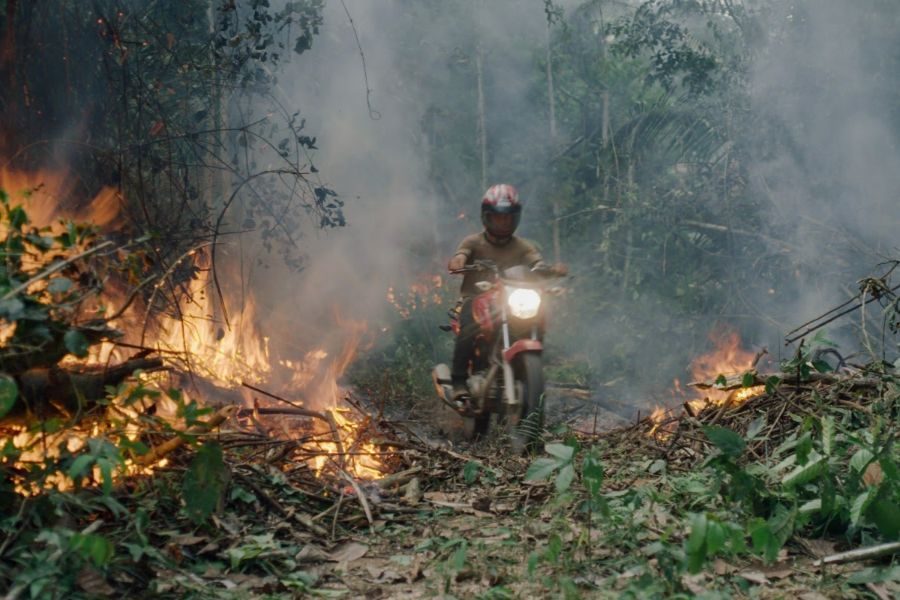 A motorcycle drives toward the camera, through the flames and smoke of the burning rainforest around them.