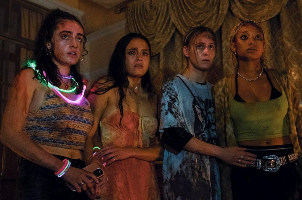 Four young women, dressed for a party but marked with dirt and blood, one still decorated with glowsticks, stand together looking scared at something unseen in front of them. 