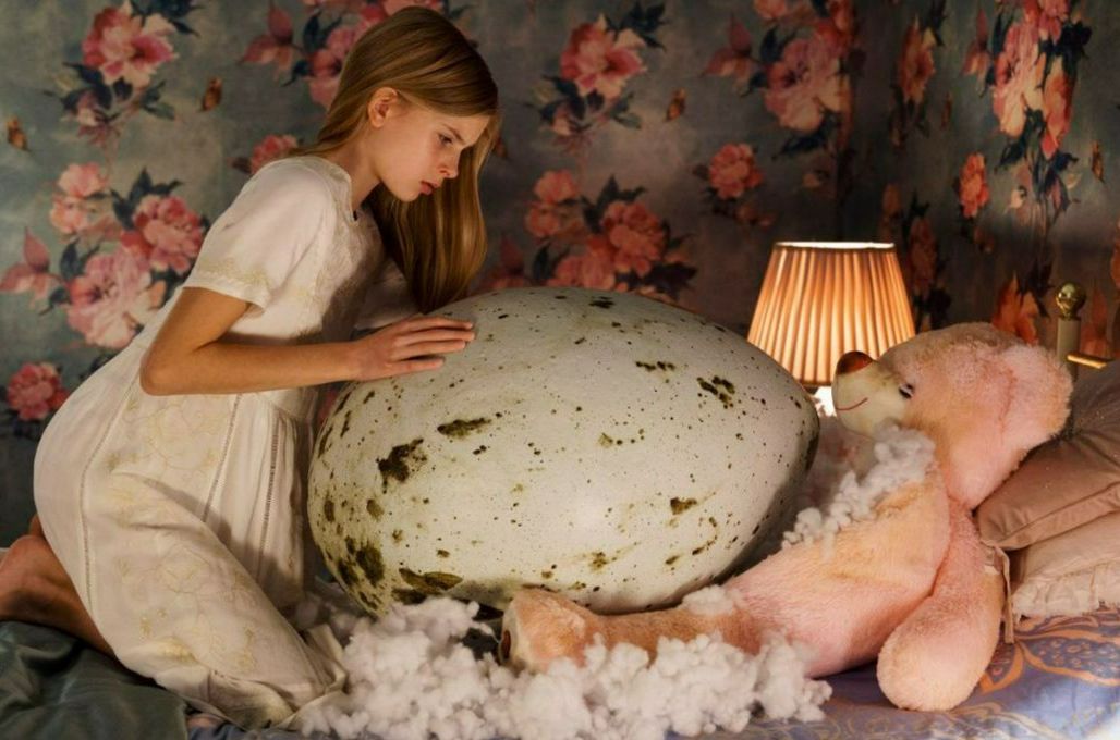 A young girl strokes a giant egg, nested in the torn up stomach of a pink teddy bear that is lying on her bed. 