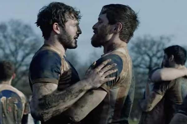 Two male rugby players, streaked with mud, embrace tenderly on the pitch. 