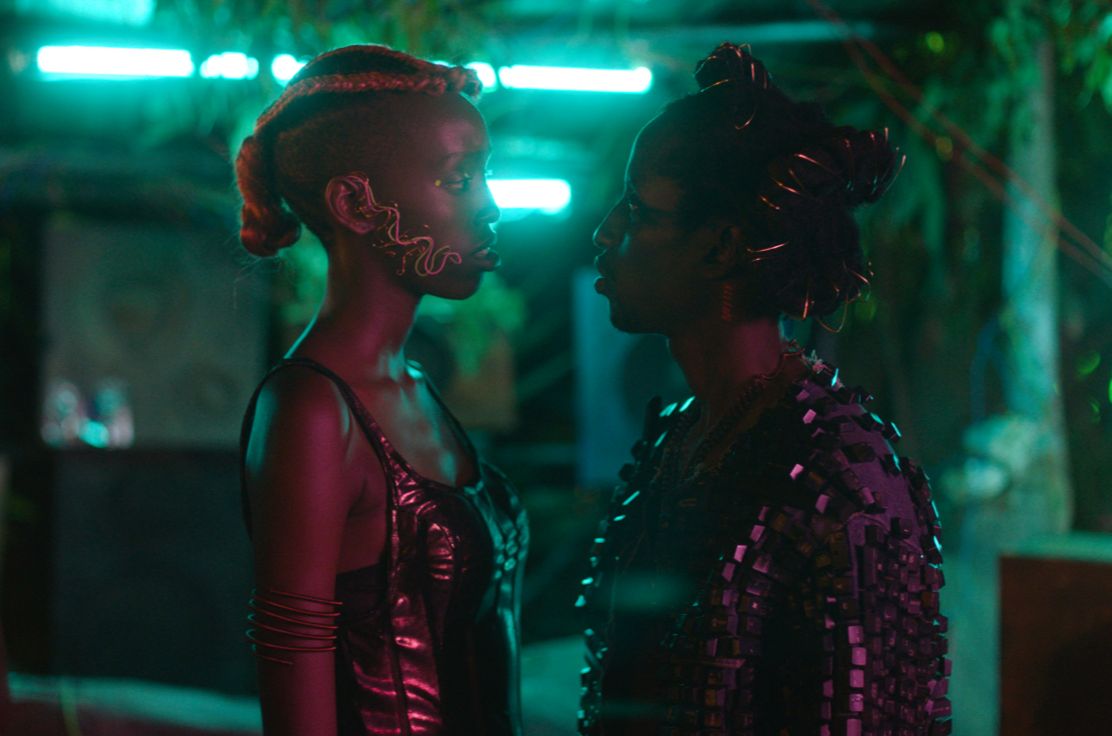 Two woman in afro-futuristic clothing stare at each other intensely, behind them the room is bathed in blue light. 