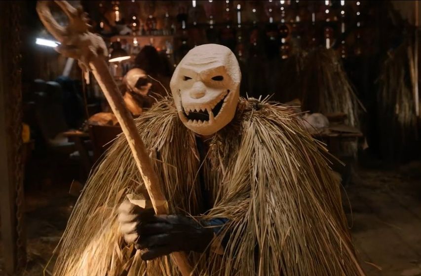 A person in ape-like skill mask and outfit covered in straw, holds an ornate staff in an old wooden shack. 