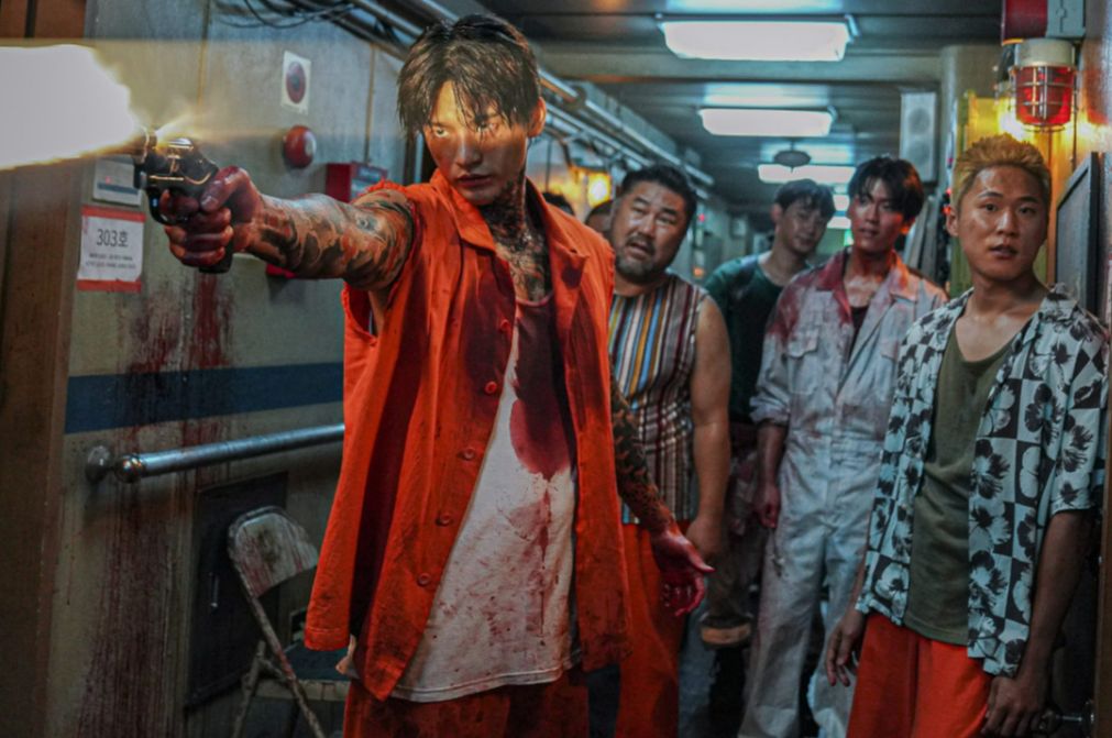A group of man, looking tough and covered in blood stand in an equally blood corridor. The man in the front wears an orange prison outfit and is firing a gun to something in the distance. 
