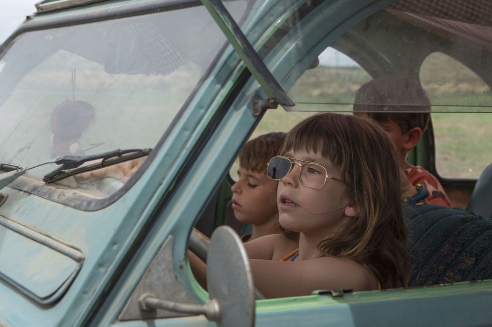 A young girl sits in the driver's seat of a beat-up old car, full of other young children.