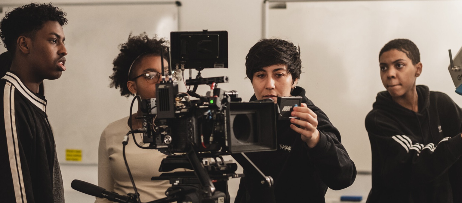 A group of young people study the screen of a large piece of camera equipment.