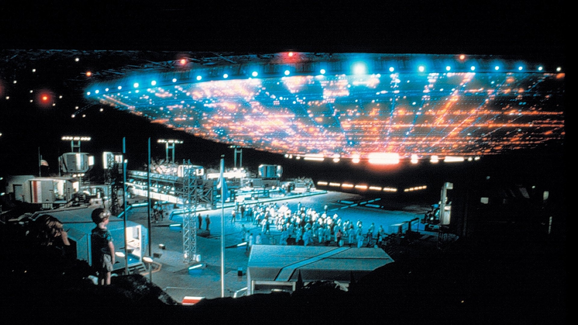A screenshot from Close Encounters of the Third Kind showing a large spaceship floating over a government base as a young child watches from nearby.