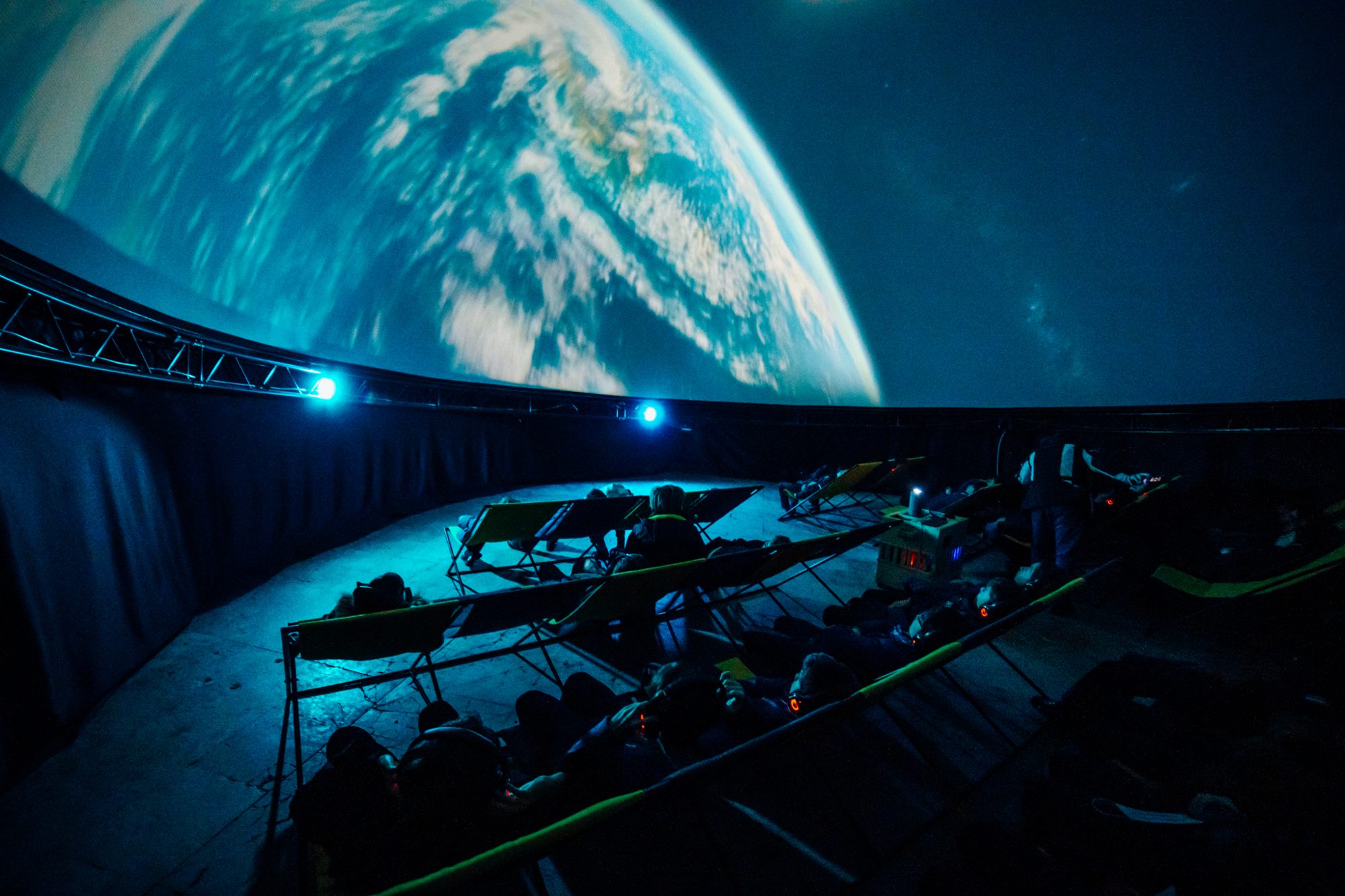 A large image of earth swirls on the domed ceiling of a planetarium, with rows of people sat back and watching show.
