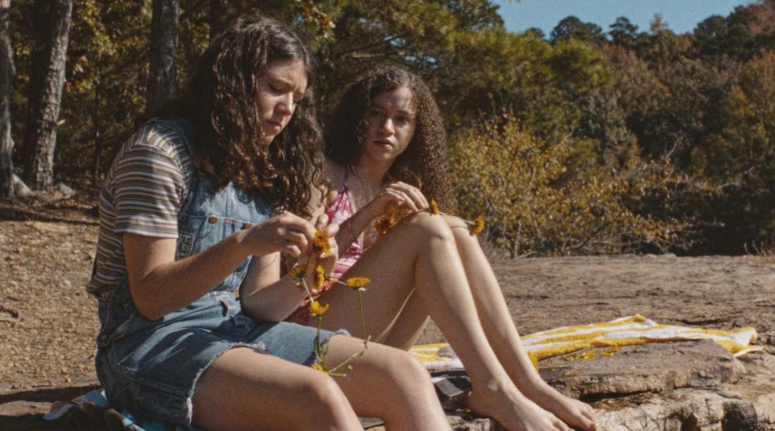 Two teenage girls sit in a clearing by a forest, making flower necklaces.