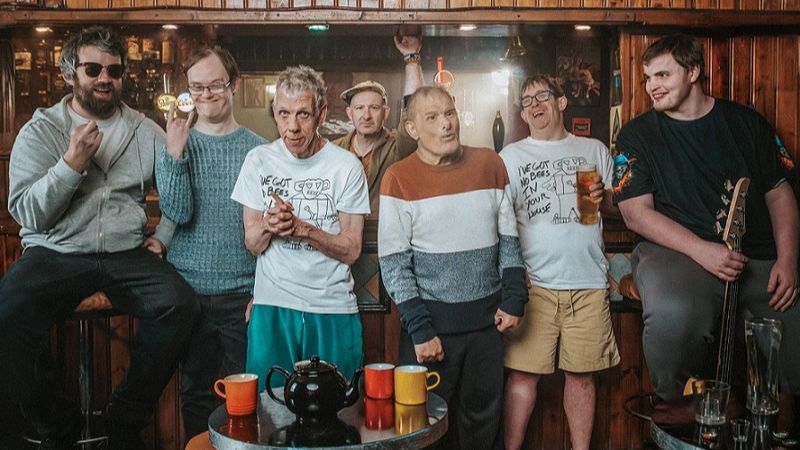 A group of people of mixed age, celebrate in a local pub, one of them holding a guitar.