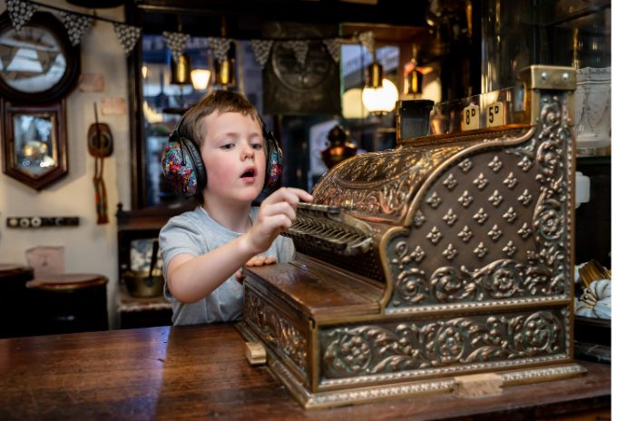 A child wearing ear defenders interacts with an old fashioned shop till