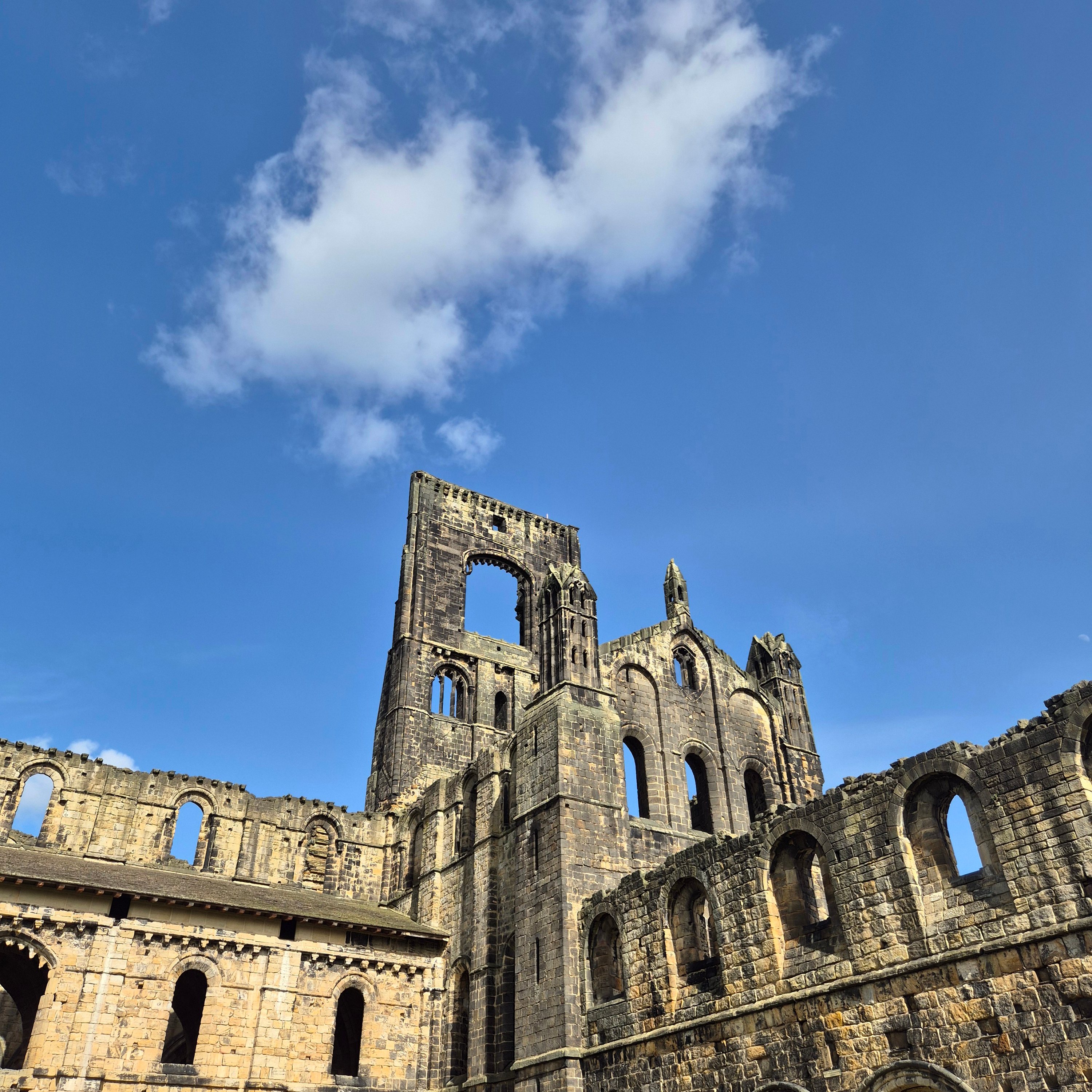 Looking up at Kirskstall Abbey ruins with a blue sky and single white cloud above
