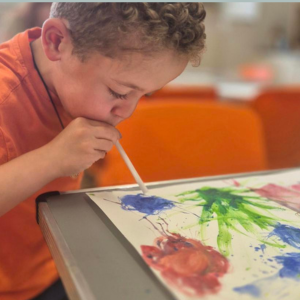 A young child in an orange top blows through a straw onto his colourful painting