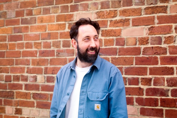 Architect David Pogson in a blue shirt and a white T-shirt stood in front of a brick wall.