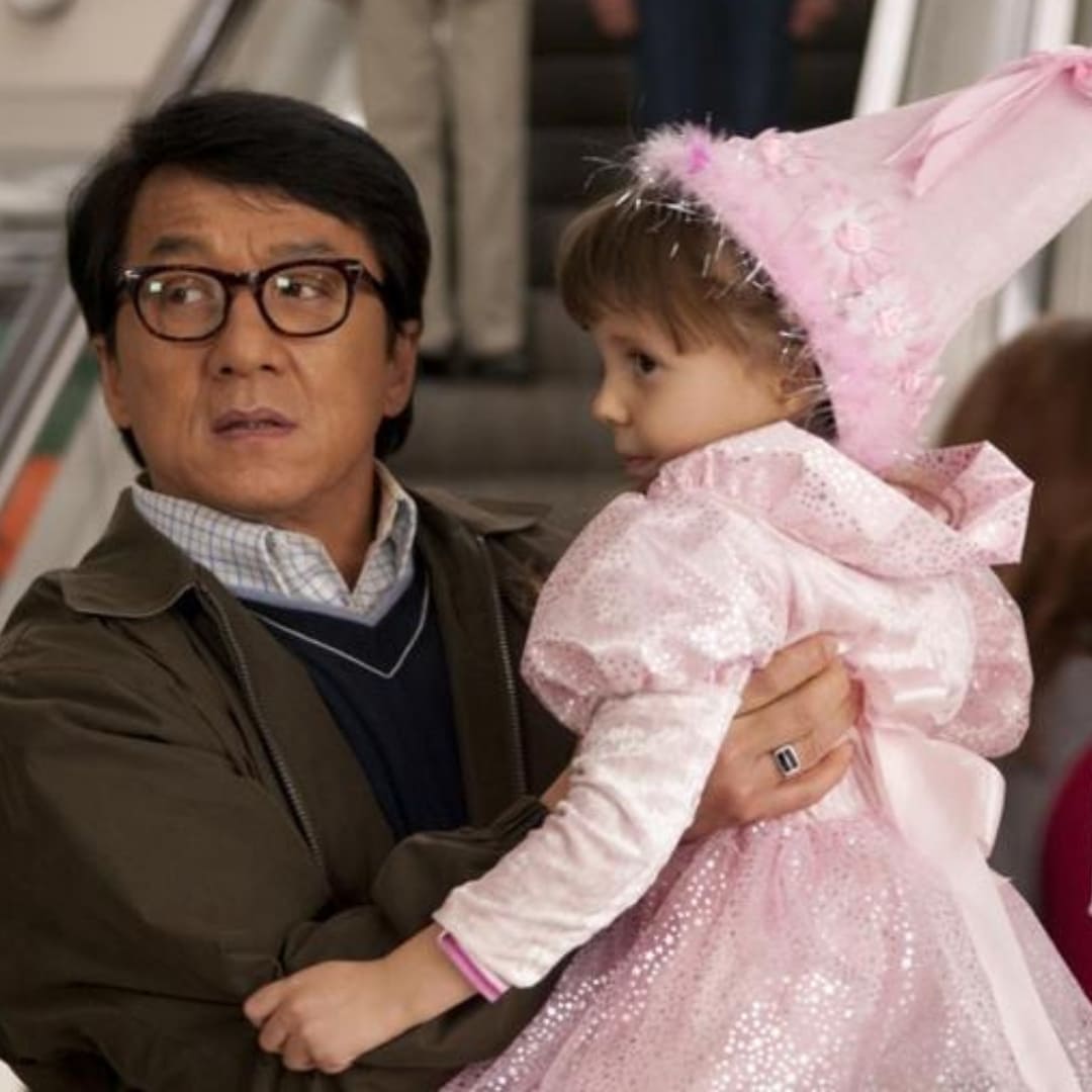 Jackie Chan holds up a small child wearing a pink princess costume: a long sleeves dress and pointed hat.