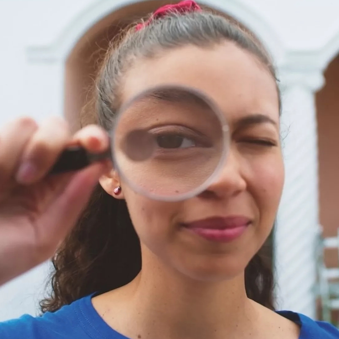 A teenage girl looks into the camera using a magnifying glass.