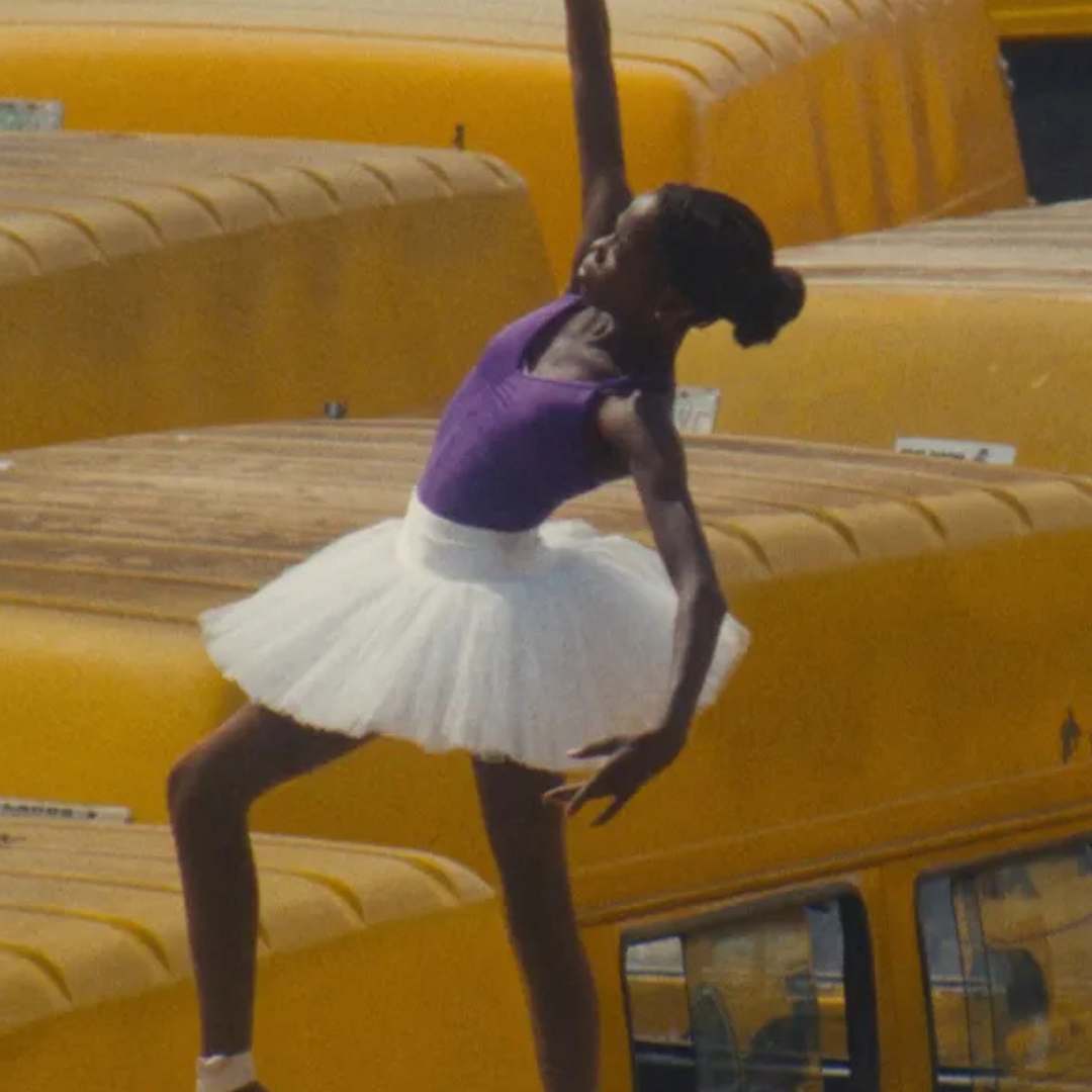 A female dancer stands on the roof of a yellow bus wearing a white tutu