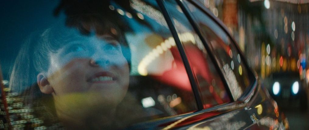 An animated depiction of Frida Kahlo sits in a wheelchair, painting a still life onto a canvas. She is wearing a floral headpiece and surrounded by plants, fruit and paintbrushes.