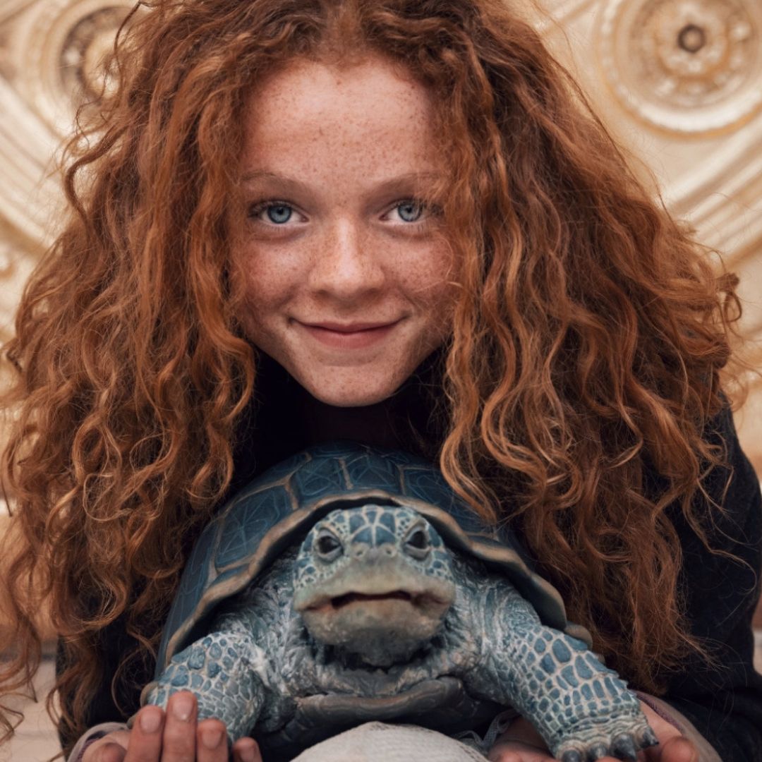 A young girl with unruly ginger hair holds a turtle and looks towards the camera.