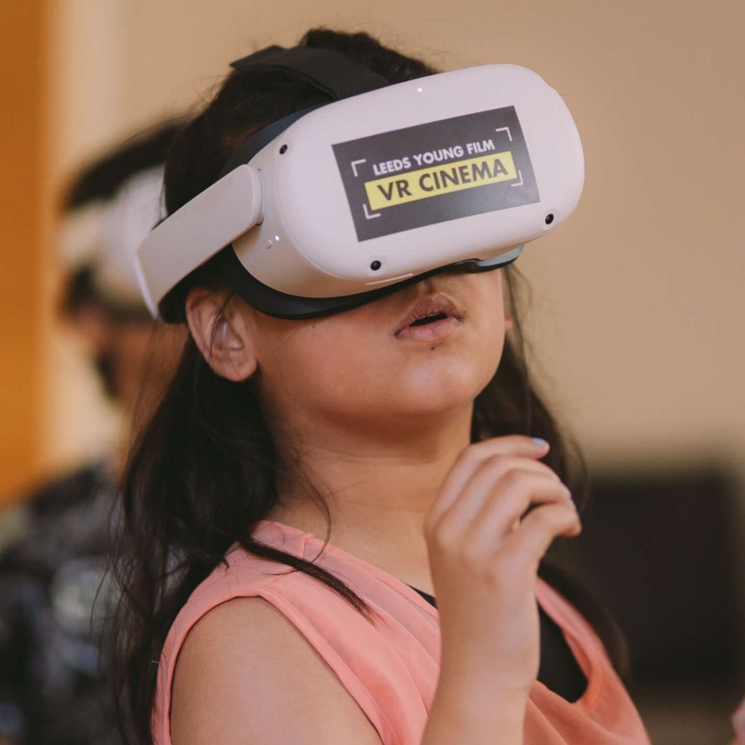 A young girl with dark hair and a pink vest wears a 'Leeds Young Film VR Cinema' VR headset.