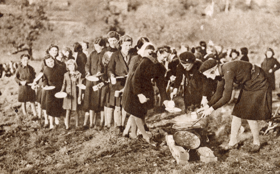 Black and white photograph showing a large group of girl guides. They are serving hot drinks from a makeshift outdoor stove