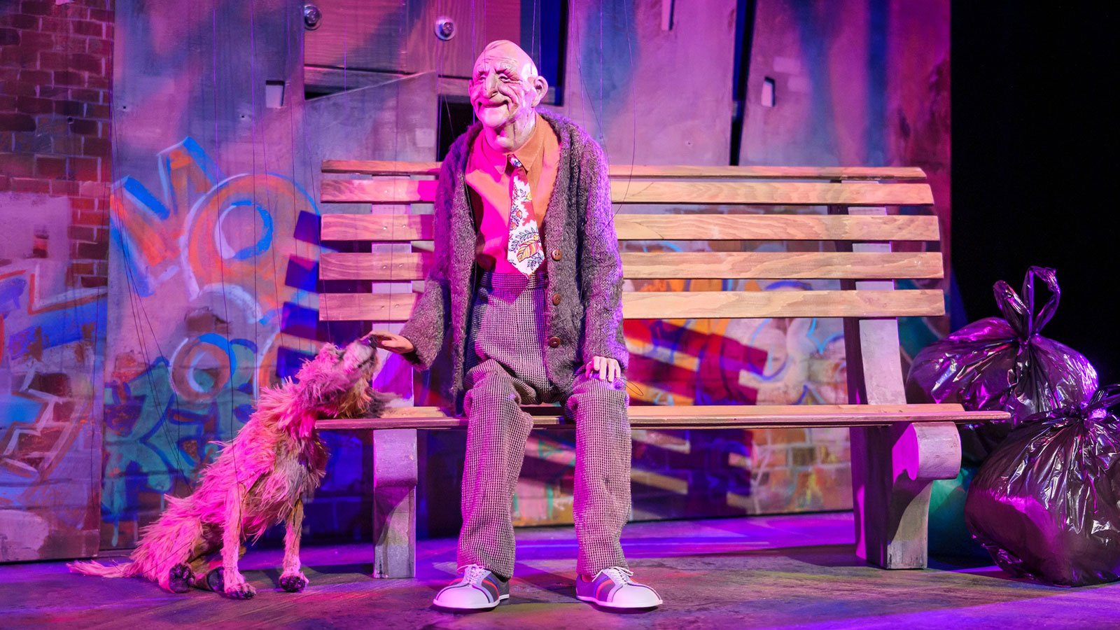 A puppet of an old man standing next to a wooden bench and a dog